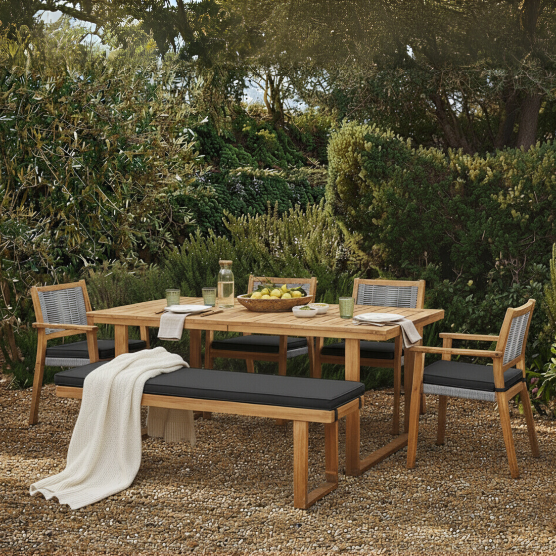 Albian 7 Seater Hardwood Outdoor Dining Set in Light Grey features a wooden table, four armchairs, and a bench with black cushions, set on a gravel patio with a lush green backdrop.