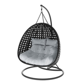 Buena 2 Person Wicker Outdoor Hanging Egg Chair in Black, features a freestanding rattan shell suspended by chains, with a round metal base and curved arm, housing thick tufted cushions for comfort.