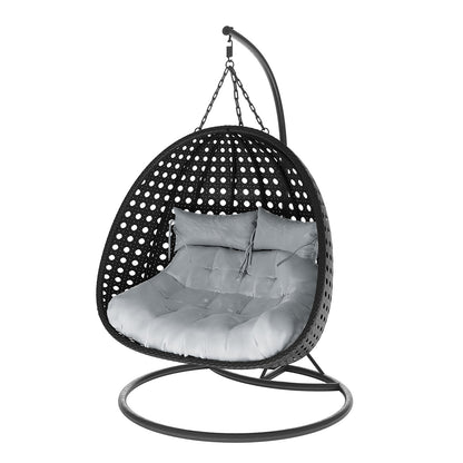Buena 2 Person Wicker Outdoor Hanging Egg Chair in Black, features a freestanding rattan shell suspended by chains, with a round metal base and curved arm, housing thick tufted cushions for comfort.