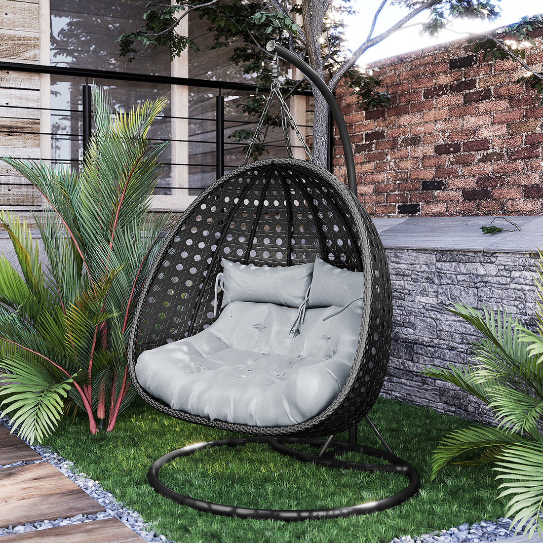 Buena 2 Person Wicker Outdoor Hanging Egg Chair in Black, featuring a tufted gray cushion and lumbar pillow, suspended from a metal stand amidst a garden setting with tropical plants.