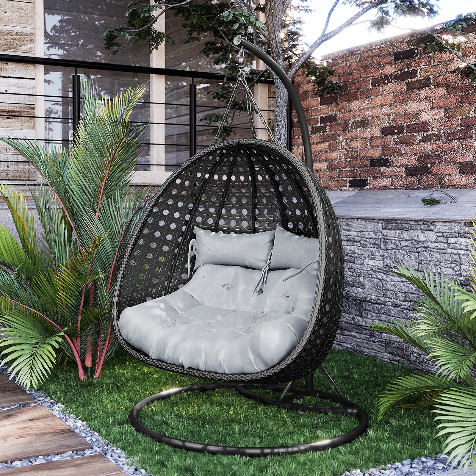 Buena 2 Person Wicker Outdoor Hanging Egg Chair in Black, featuring a tufted gray cushion and lumbar pillow, suspended from a metal stand amidst a garden setting with tropical plants.