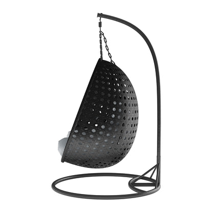 Buena 2 Person Wicker Outdoor Hanging Egg Chair in Black features a perforated half-shell seat with cushions, suspended by chains from a curved metal arm, perfect for relaxing outdoors.