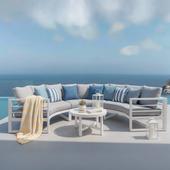 Callanna 5-Seater Round Outdoor Lounge Set in White showcasing a curved sectional sofa with pillows, a striped throw, and a white coffee table with a succulent, books, and lanterns, overlooking an ocean view.