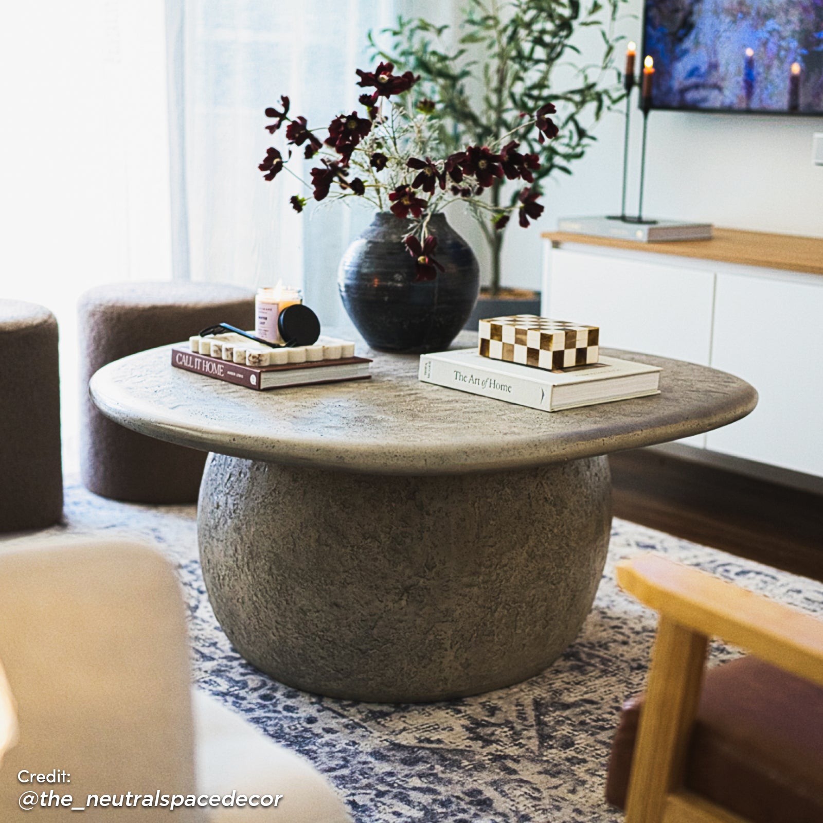 Cam Indoor & Outdoor Round Concrete Coffee Table - Stone, showcasing its round shape and textured concrete finish, situated on a coffee table with a vase, houseplant, and indoor furniture elements around it.