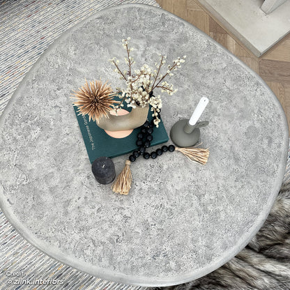 Cam Indoor & Outdoor Round Concrete Coffee Table - Stone, featuring a round, textured concrete surface, ideal for modern farmhouse aesthetics, shown on a table with decorative objects, exemplifying its spacious, durable design.