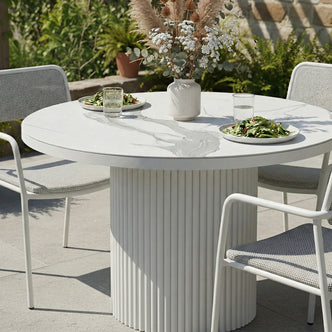 Round outdoor dining table with a white marbled stone top and a white fluted pedestal base, paired with matching mesh-backed chairs on a sunny patio.