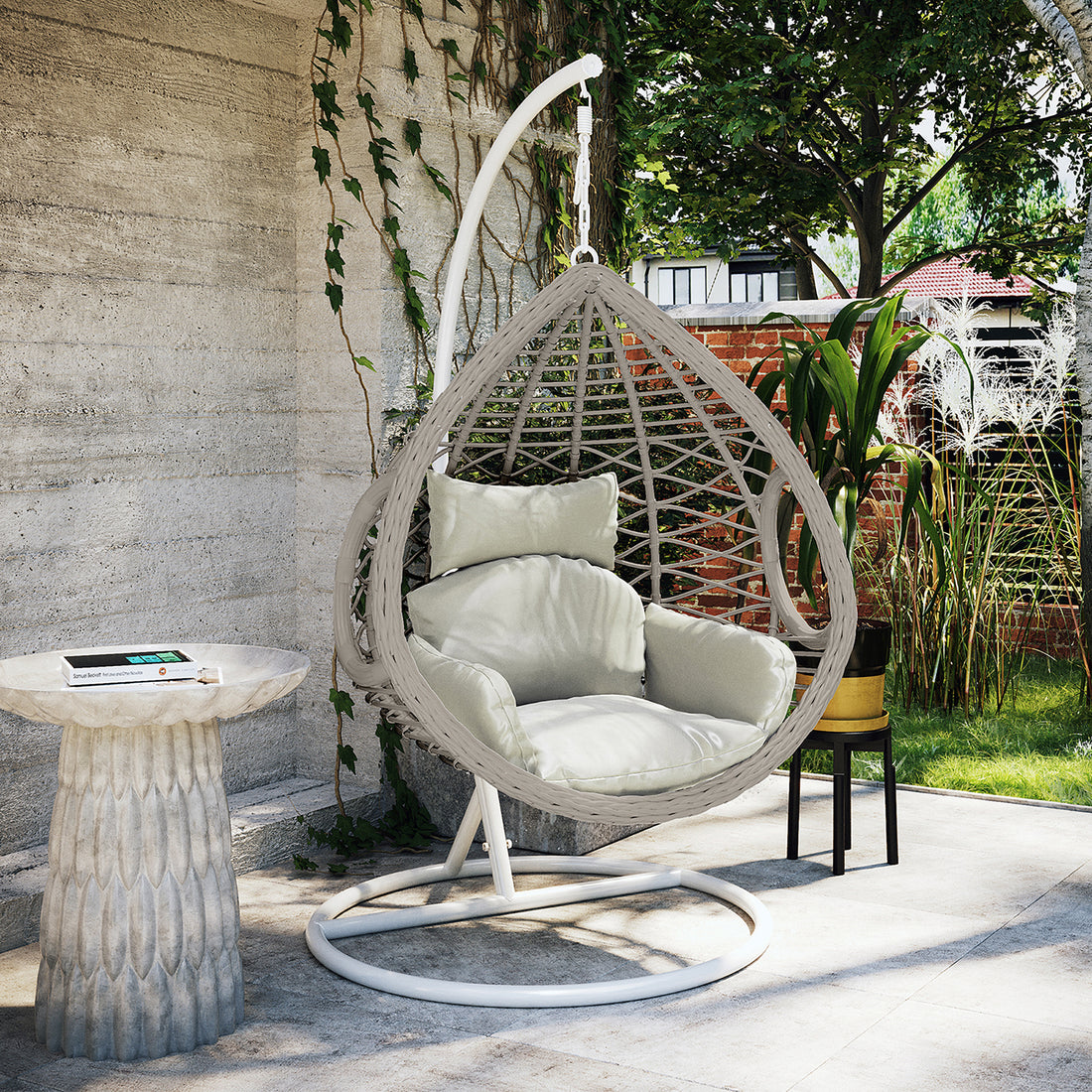 Hilda Large Outdoor Hanging Egg Chair & Cushion in Sage, teardrop-shaped with plush cushions, suspended from a white stand, alongside a small pedestal table and potted plant in a garden setting.