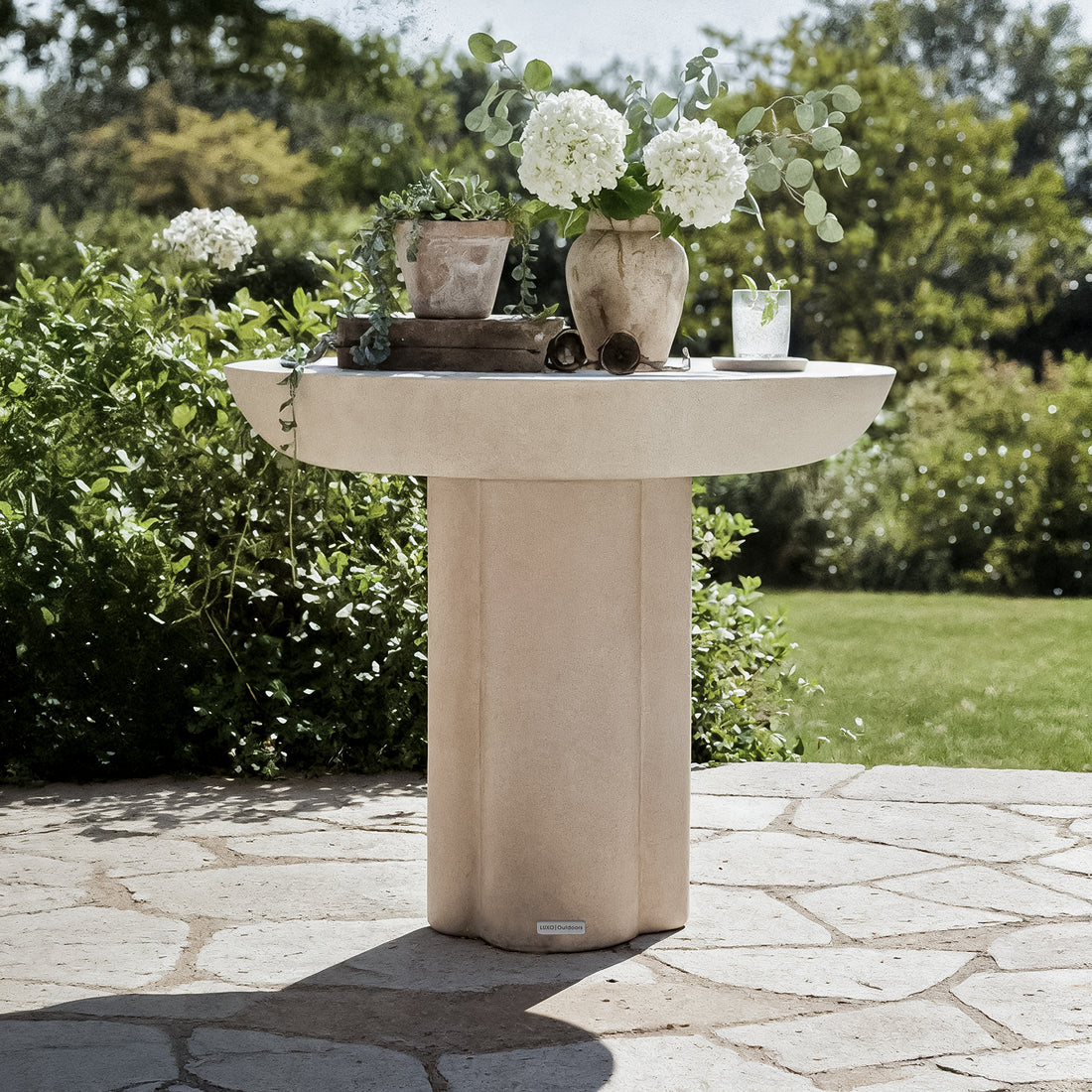 Jareth Round Concrete Outdoor Entry Table in Ivory on a patio, displaying a clay pot with succulents, a vase with hydrangeas and eucalyptus, and a glass with ice and mint.