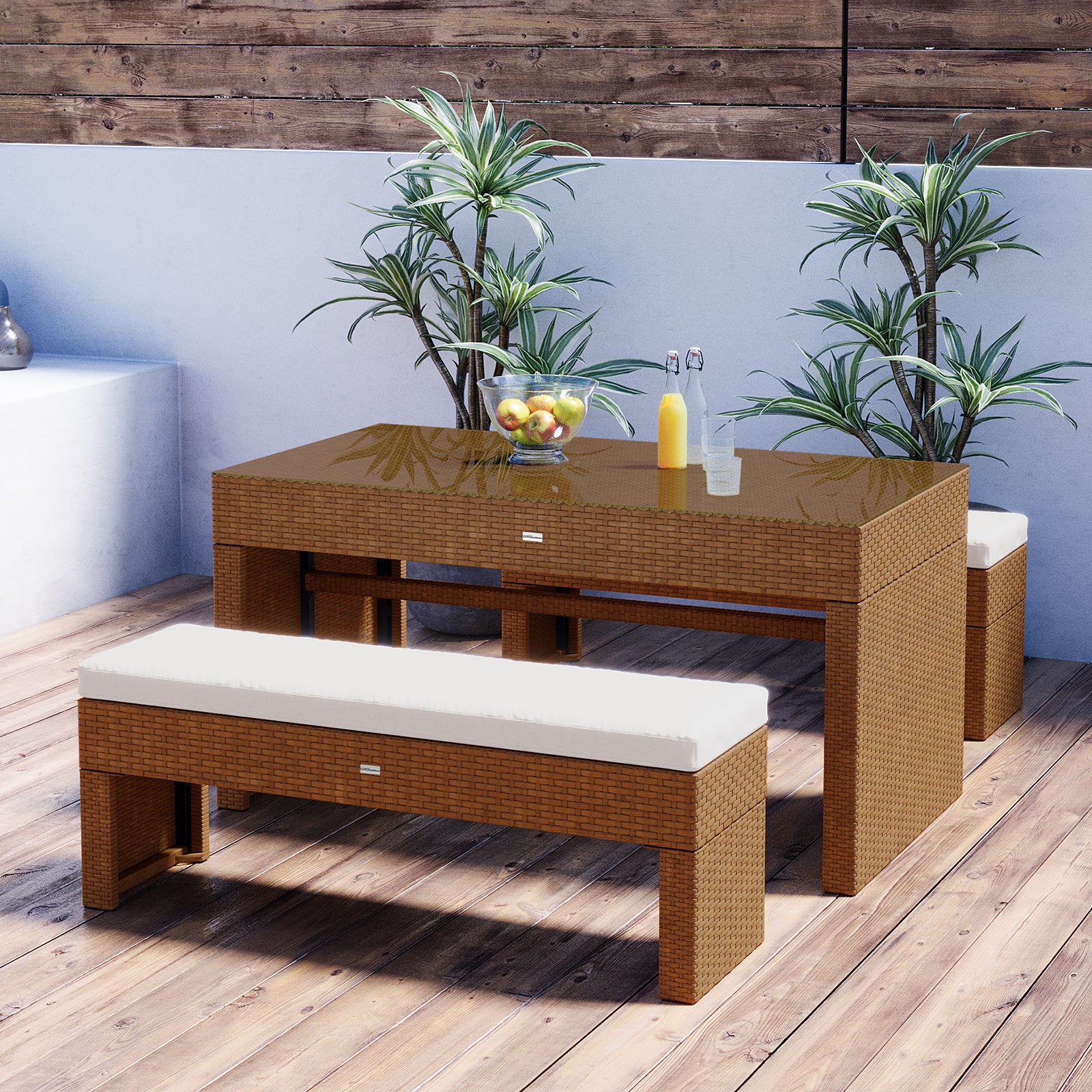 Kummi 4 Seater Outdoor Dining Bench Set features a wicker table with a glass top, two matching benches with cushions, a fruit bowl, and potted plants, creating a stylish outdoor setup.
