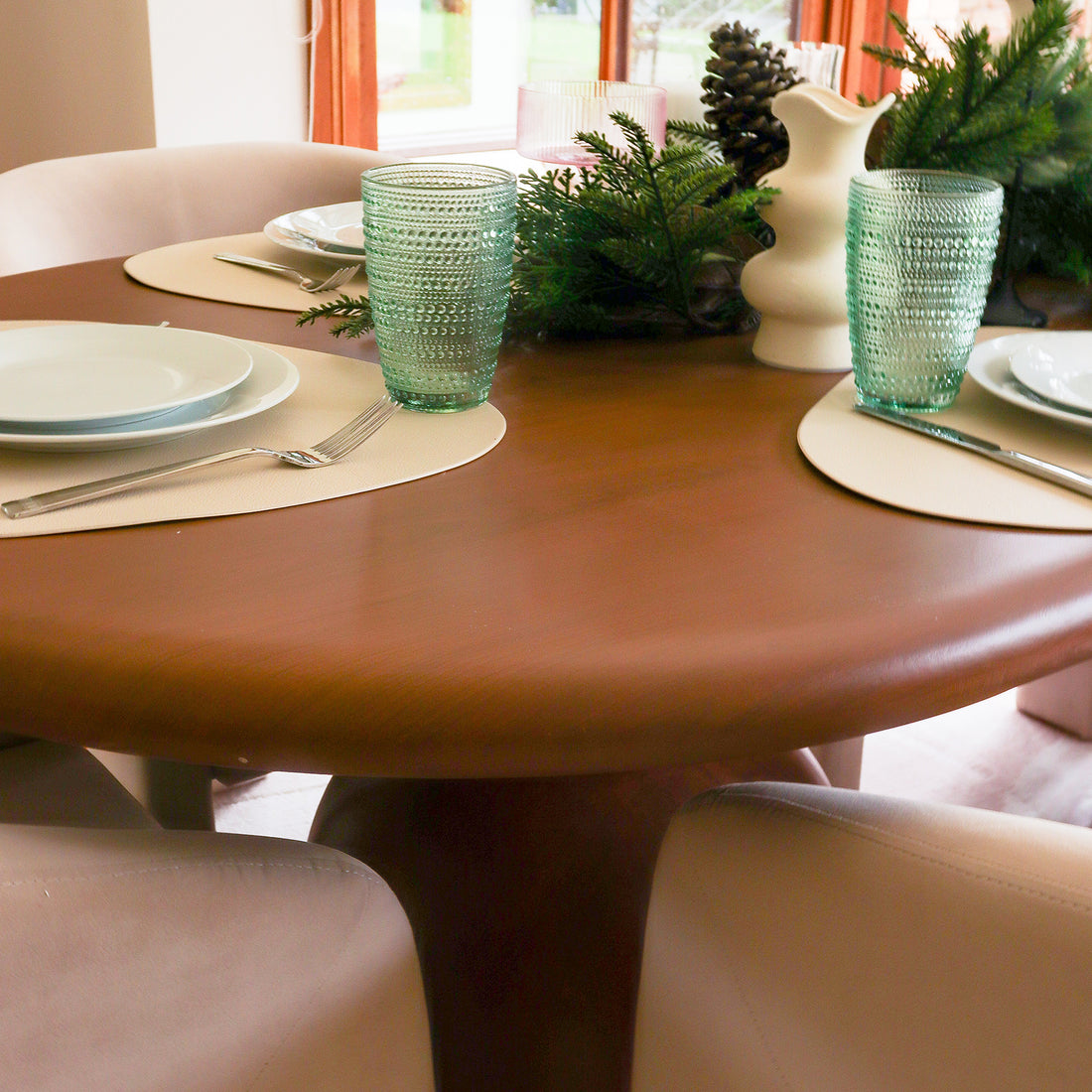 Close-up of the Lumen 220cm Oval Outdoor Concrete Dining Table in Chestnut, elegantly set with white plates, pale-green glasses, and an evergreen centerpiece, surrounded by plush cream-colored chairs.