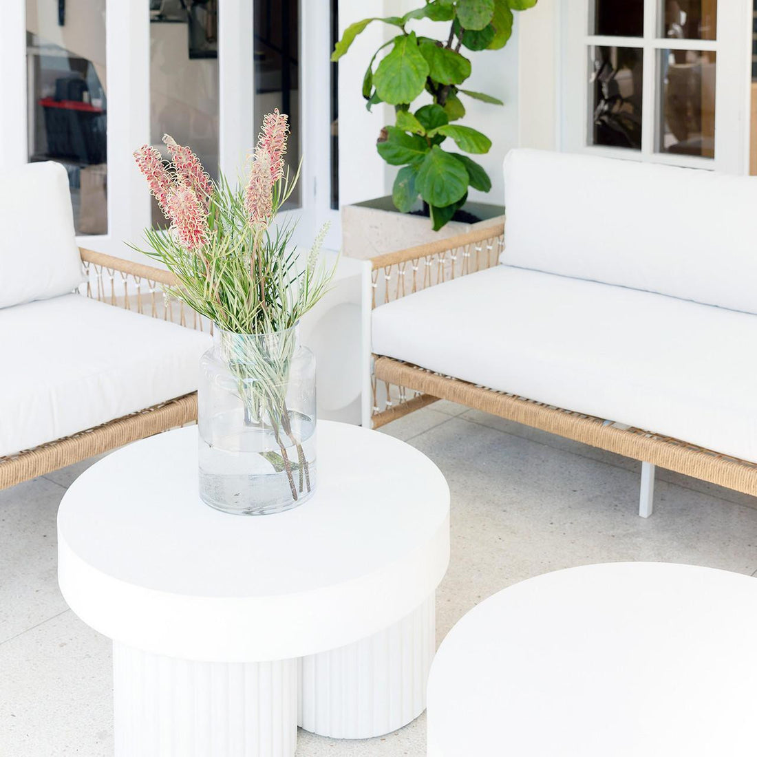Nuevo 4 Seater Wicker Outdoor Lounge Set in Light Beige, featuring white cushions and woven rattan frames, placed in a bright, minimalist nook with two round white coffee tables.