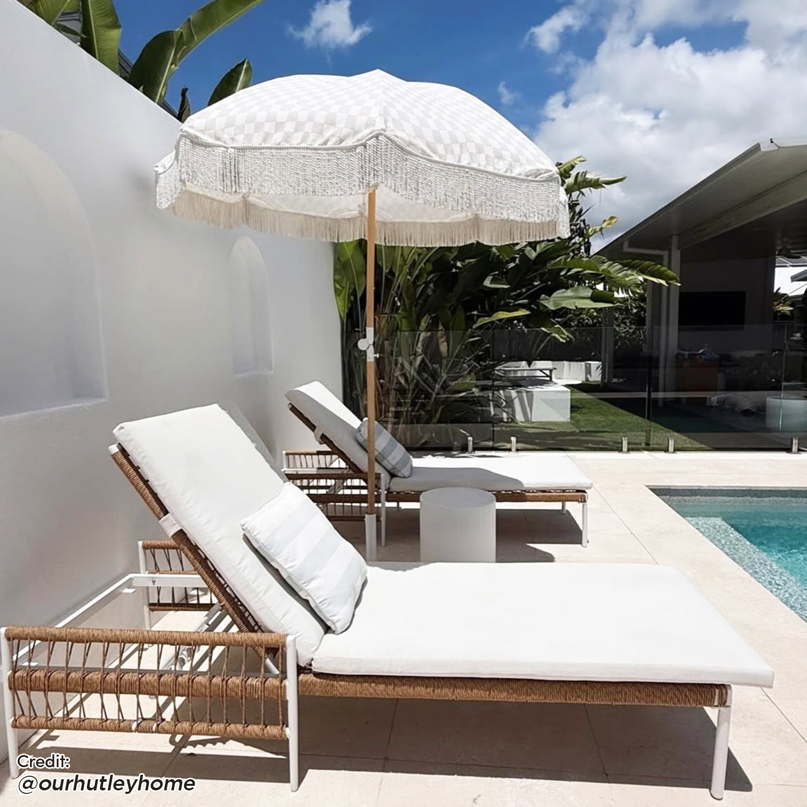 Sibutu Wicker Outdoor Sun Lounger in Light Beige: Two woven-rope lounge chairs with white cushions and bolster pillows, angled by a pool, accompanied by a small round side table and fringed market umbrella.