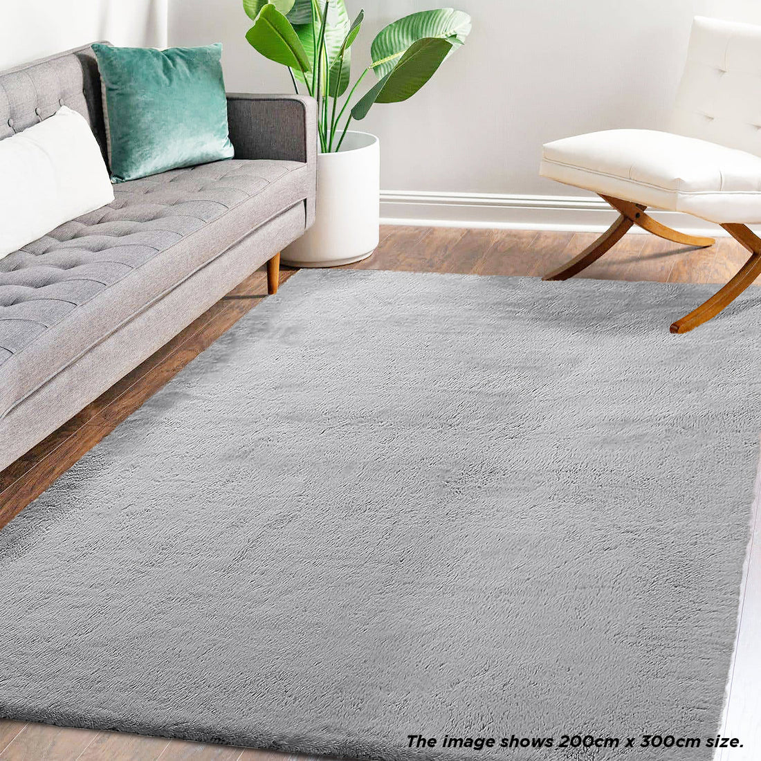 Azaliya 200x300cm Faux Rabbit Fur Floor Rug in Silver displayed in a modern living room with a mid-century gray sofa, white accent chair, and potted plant on a warm wood-tone floor.