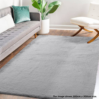 Azaliya 200x300cm Faux Rabbit Fur Floor Rug in Silver displayed in a modern living room with a mid-century gray sofa, white accent chair, and potted plant on a warm wood-tone floor.