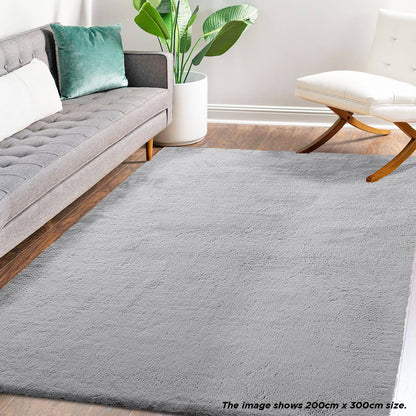 Azaliya 200x300cm Faux Rabbit Fur Floor Rug in Silver displayed in a modern living room with a mid-century gray sofa, white accent chair, and potted plant on a warm wood-tone floor.