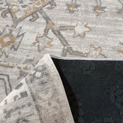 Bortan Persian Indoor Rug 120x180cm features a light grey pile with muted gold-and-charcoal abstract motifs, showcasing a flipped corner revealing its black, dotted non-slip backing.