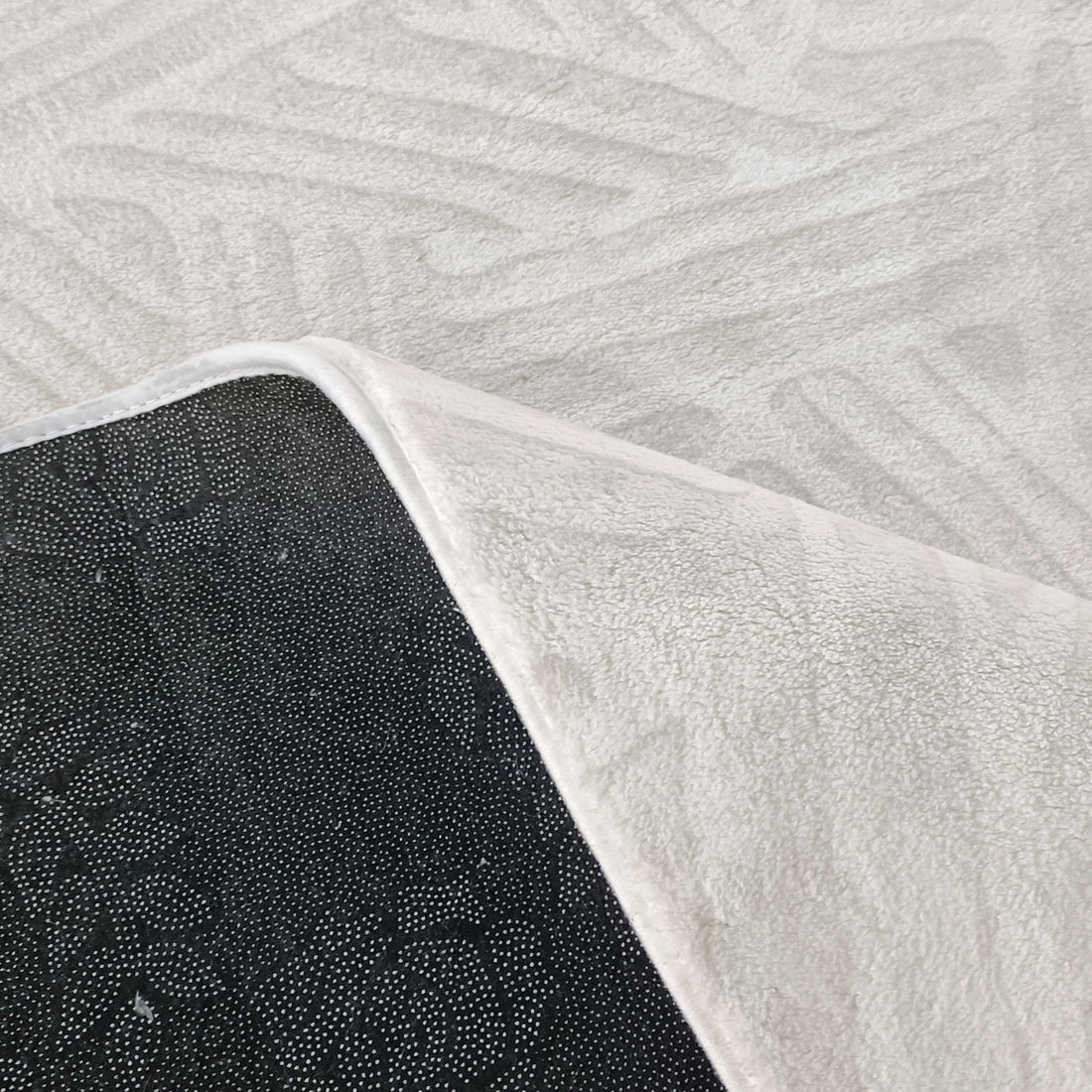 Close-up of Malung 200x300cm Contemporary Indoor Floor Rug, featuring plush off-white texture with a flipped corner revealing black dot-patterned non-slip backing on a light patterned floor.