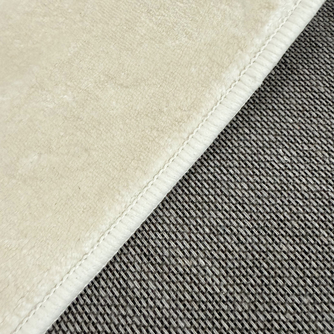 Close-up of Pavla 200 x 300cm Oval Shaggy Rug in Beige, showcasing the plush cream microfiber texture meeting a gray woven textile, highlighting its soft, shaggy design and stitched seam detailing.