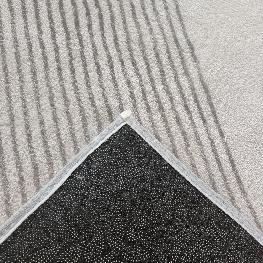 Close-up of Ramvik 200x300cm Abstract Indoor Floor Rug showing flipped corner, revealing black, dotted non-slip backing. The rug's abstract design with squiggly lines adds modern flair against a striped gray carpet.