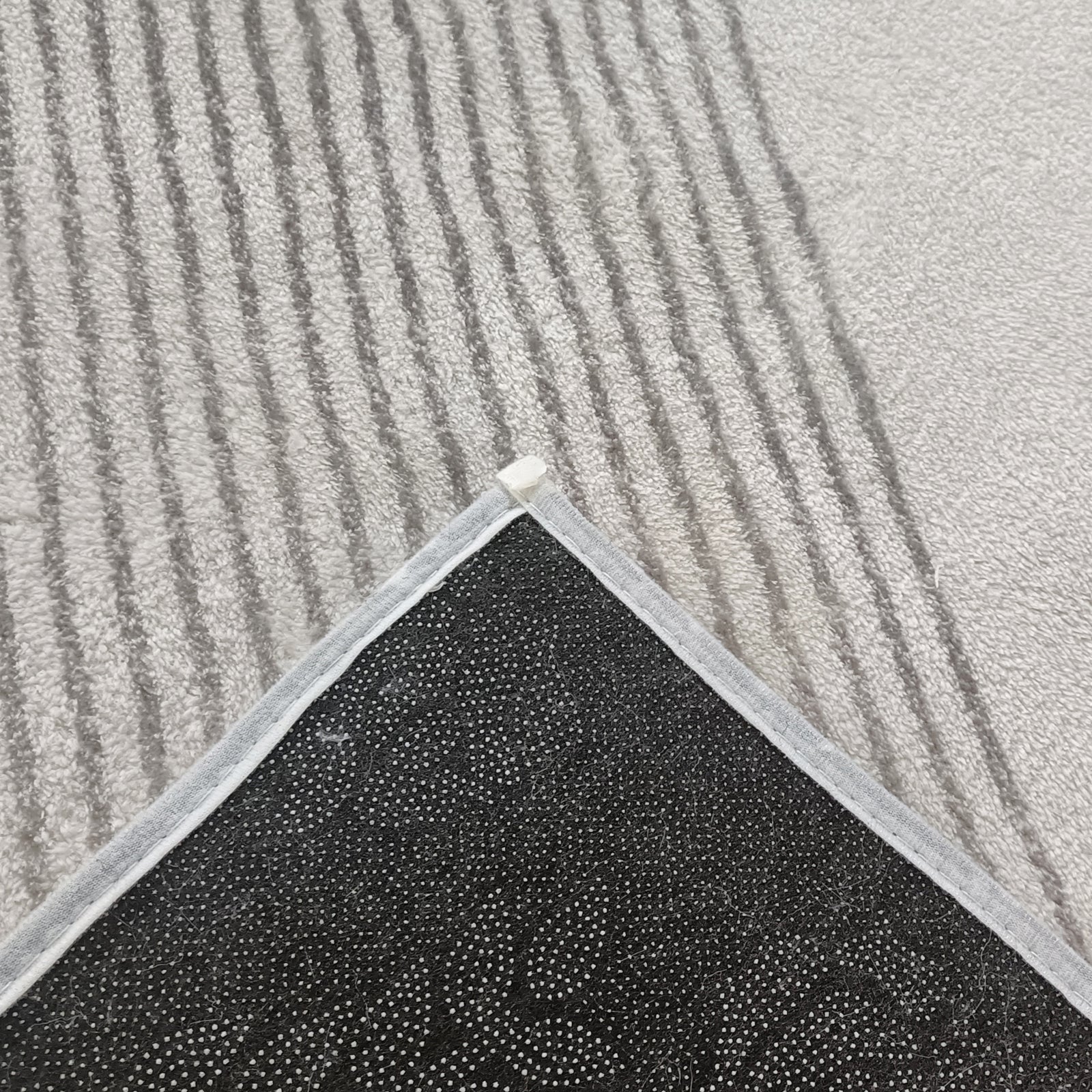 Close-up of Ramvik 200x300cm Abstract Indoor Floor Rug showing flipped corner, revealing black, dotted non-slip backing. The rug's abstract design with squiggly lines adds modern flair against a striped gray carpet.
