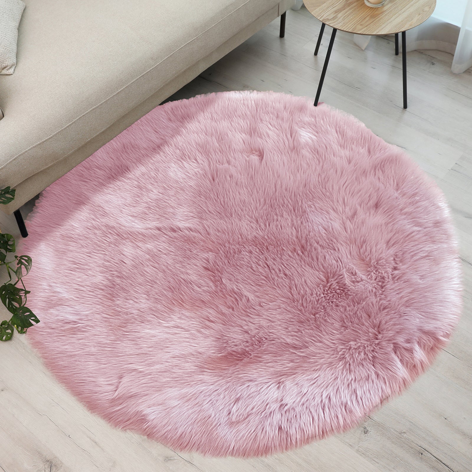 Cozy corner with a Shaddi 120cm Round Shaggy Soft Fluffy Fur Rug in Pink on light wood floor, bordered by beige sofa and wooden side table, complemented by green plant foliage.