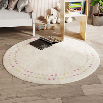 Shirin 160 Round Polka Dot Kids Indoor Rug on light wood flooring, cream with pastel dots, an open book centered. Nearby, white bed frame, wooden toy train, white teddy bears, and children's books.