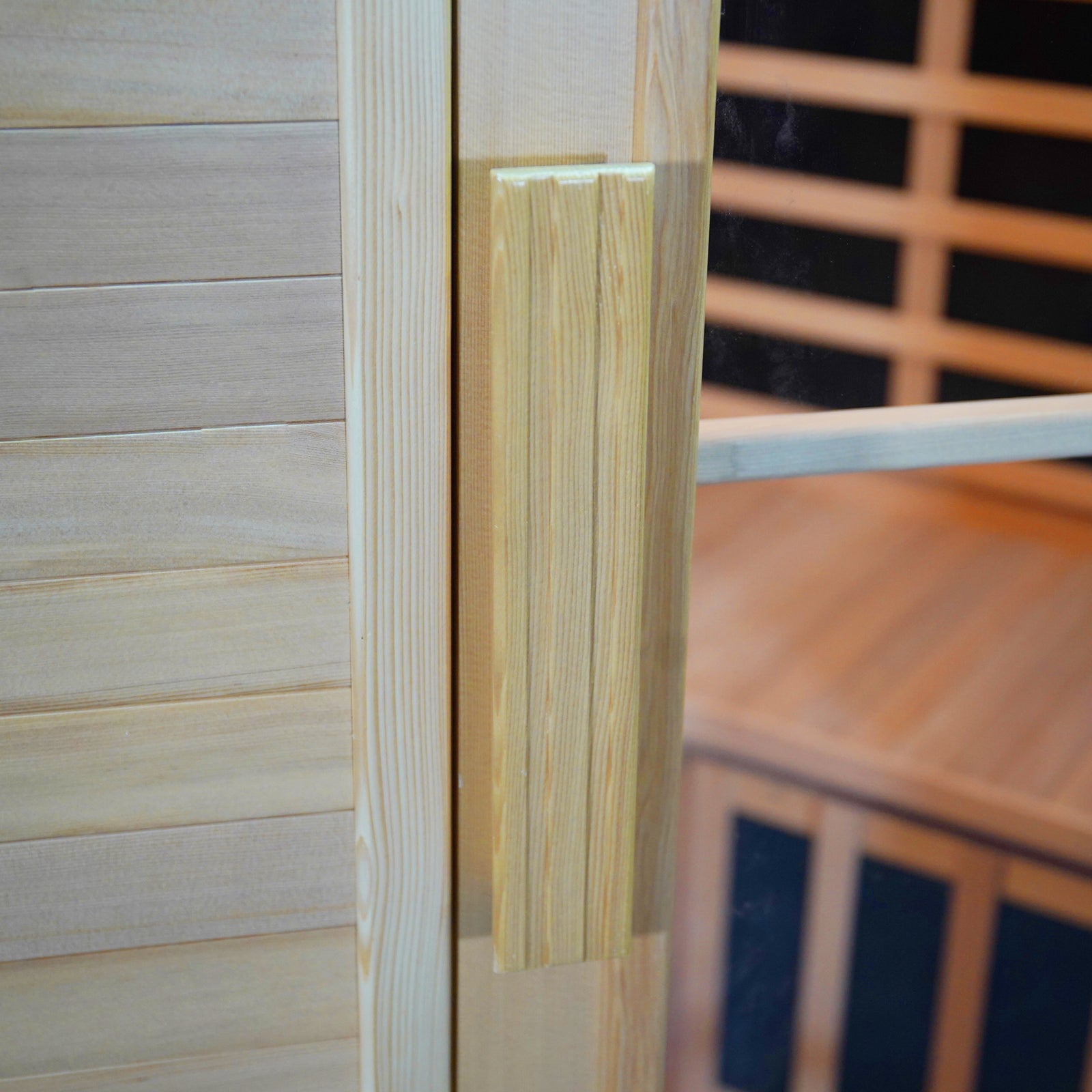 Taavi 4 Person Corner Carbon Fibre Infrared Indoor Sauna features a light-toned wooden door with a vertical wood handle on a glass panel, revealing a warm sauna interior with slatted benches.