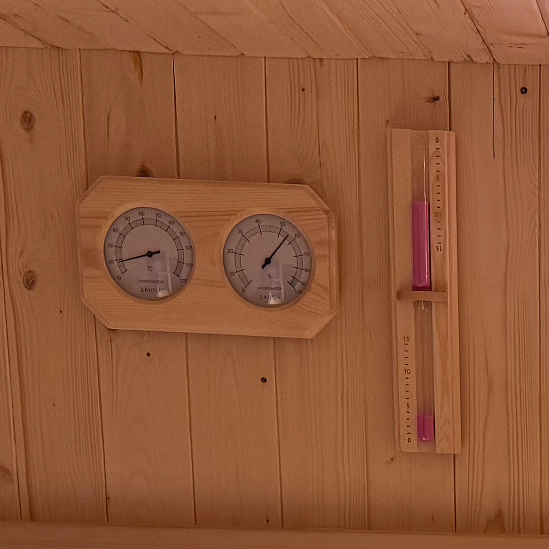 Tarni 2-Person Finnish Outdoor Sauna interior wall with a mounted thermometer and hygrometer, alongside a wooden-framed sand-glass timer with pink sand, enhancing your relaxation experience.