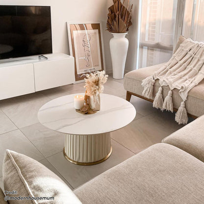 Combee 80cm Sintered Stone Coffee Table Cream White displayed with a smooth stone top and gold chromed base, enhancing a living room ambiance with a cozy white blanket and elegant glassware nearby.