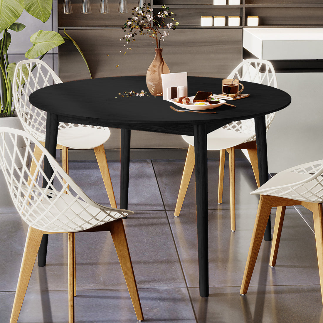 Edna 120cm Round Hardwood Dining Table in Black with a ceramic vase, pastries, and coffee on top, surrounded by four white lattice-style chairs in a modern kitchen setting.