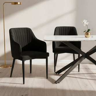 Elly Curved Velvet Dining Chairs Set of 2 in Black, featuring plush upholstery, curved high-back with armrests, and tapered legs, positioned around a marble-top table with a gold vase and orchid.