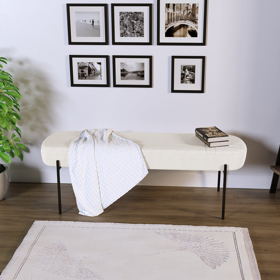 Gavin Dining Bench - Cream White, featuring sumptuous upholstery, slender black metal legs, and a soft throw, complemented by a grid of black-framed prints above and a potted plant beside.