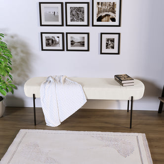 Gavin Dining Bench - Cream White, featuring sumptuous upholstery, slender black metal legs, and a soft throw, complemented by a grid of black-framed prints above and a potted plant beside.