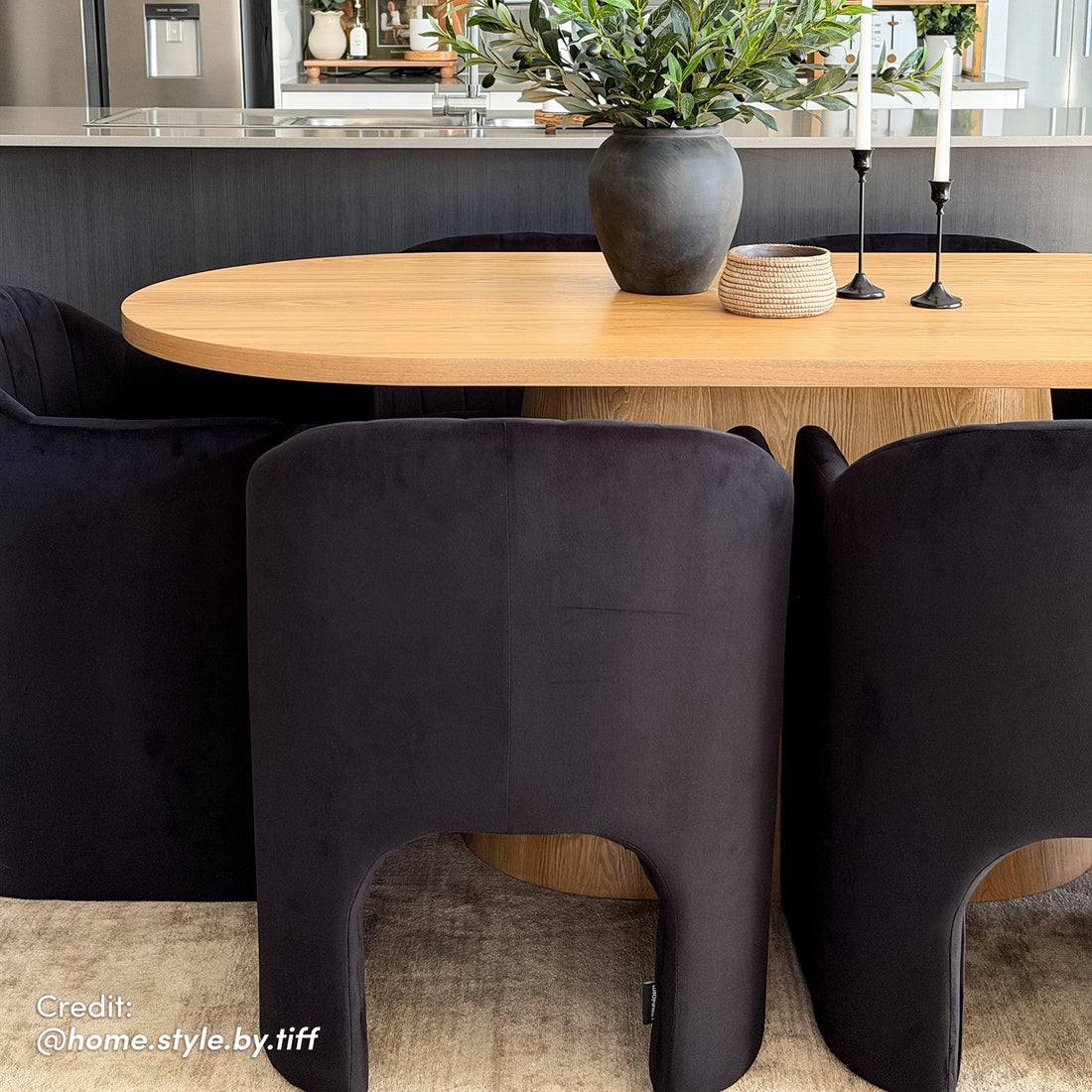 Gigi 180cm Oval Wooden Dining Table in Oak surrounded by black chairs, features a dark ceramic vase and candleholders. A modern dining space with a kitchen island visible in the background.