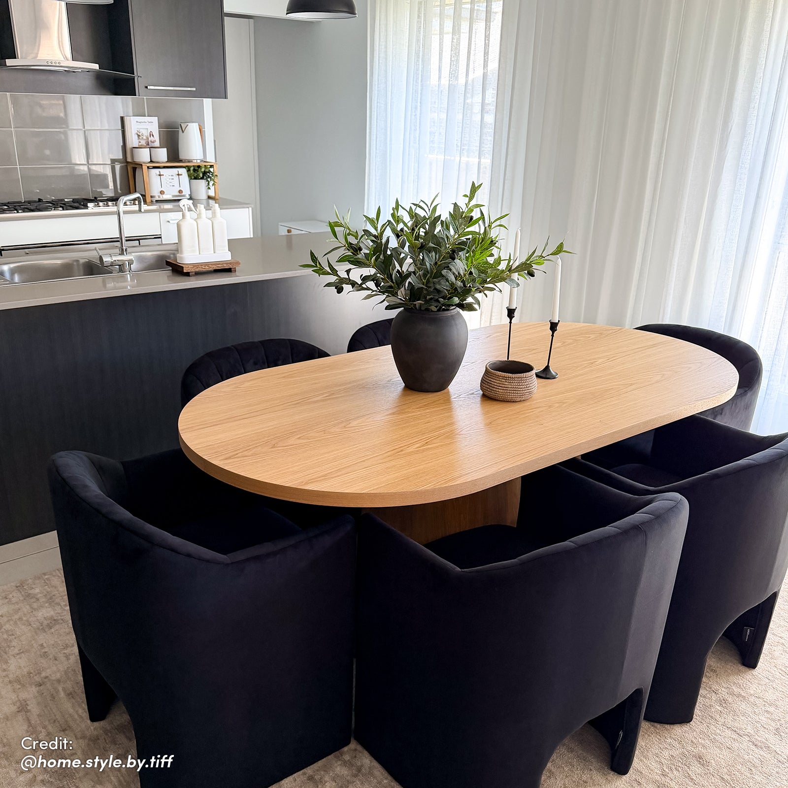 Gigi 180cm Oval Wooden Dining Table in Oak, elegantly styled with a matte-black vase, candleholders, and basket, harmonizes in a bright, minimalist dining-kitchen setting with black chairs.