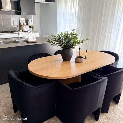 Gigi 180cm Oval Wooden Dining Table in Oak, elegantly styled with a matte-black vase, candleholders, and basket, harmonizes in a bright, minimalist dining-kitchen setting with black chairs.