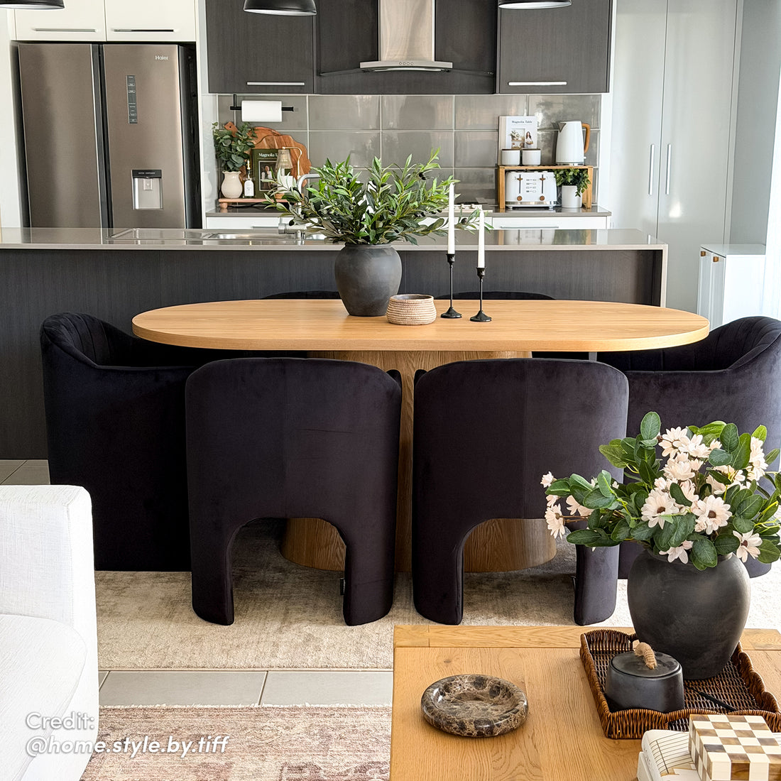 Gigi 180cm Oval Wooden Dining Table in Oak, featuring a light wood finish, surrounded by six black-velvet chairs. Adorned with a ceramic vase, basket, and candle holders, exemplifying modern elegance.