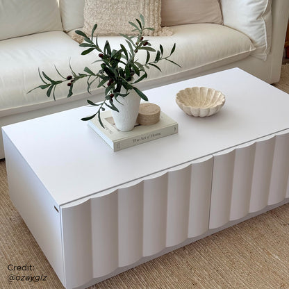 Hamish 120cm Coffee Table with Storage featuring a modern wave design, visible drawers, and a spacious top, complemented by indoor decor elements like a plant in a vase on the table.