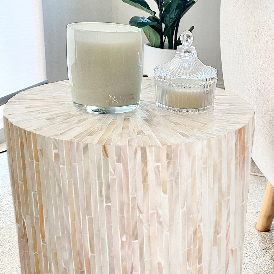 Lulu Mother of Pearl Inlay Side Table in Cream with shimmering mosaic surface, topped with two candles and a small potted plant, adjacent to a light-colored upholstered chair.