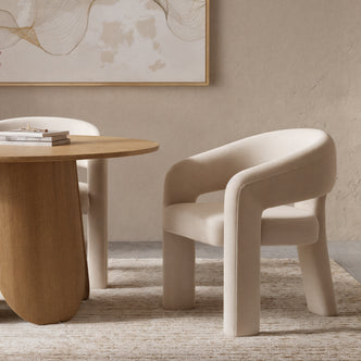 Mariah 3 Legged Velvet Accent Chair in Beige with thick, rounded legs and a gently curved backrest, placed on a textured rug beside a round wooden table in a serene, minimalist setting.