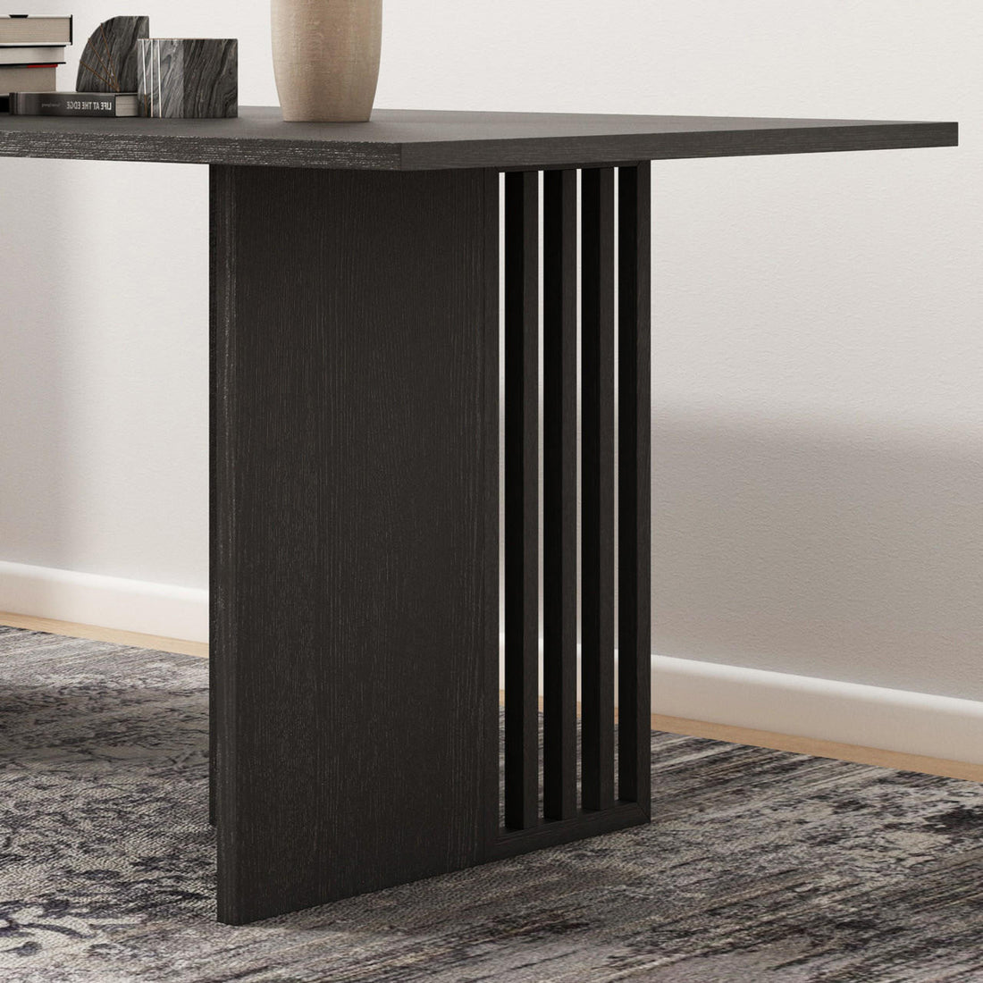 Close-up of the Rowley 180cm Dining Table Black, showcasing one modern, dark-stained wooden leg with inset vertical slats, set on an abstract-patterned rug, with books and a vase on the tabletop.