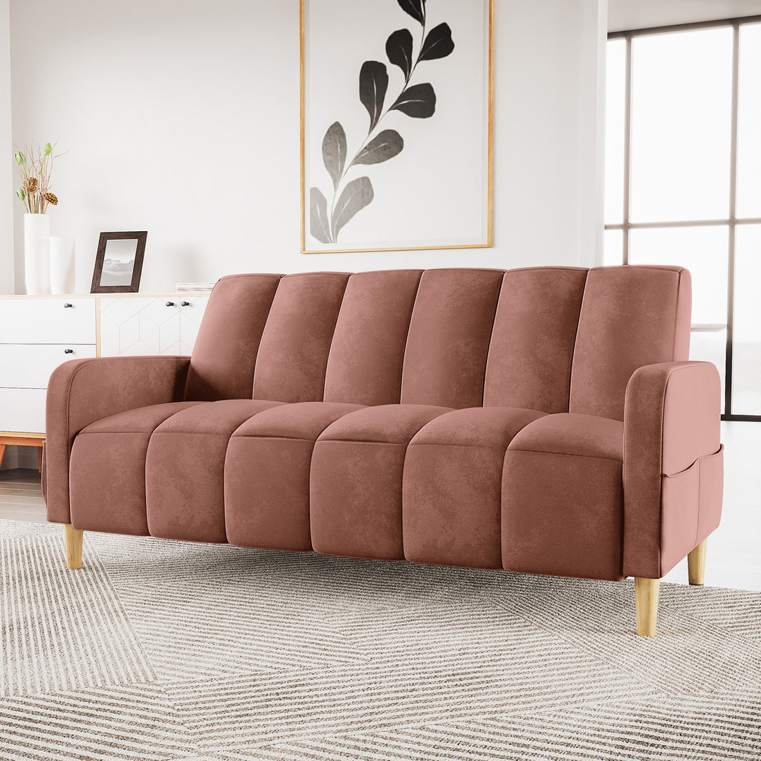Anica 3 Seater Velvet Sofa Bed in Blush with vertical channel tufting, rose-toned upholstery, and light wood legs, set on a geometric rug in a bright, minimalist room.