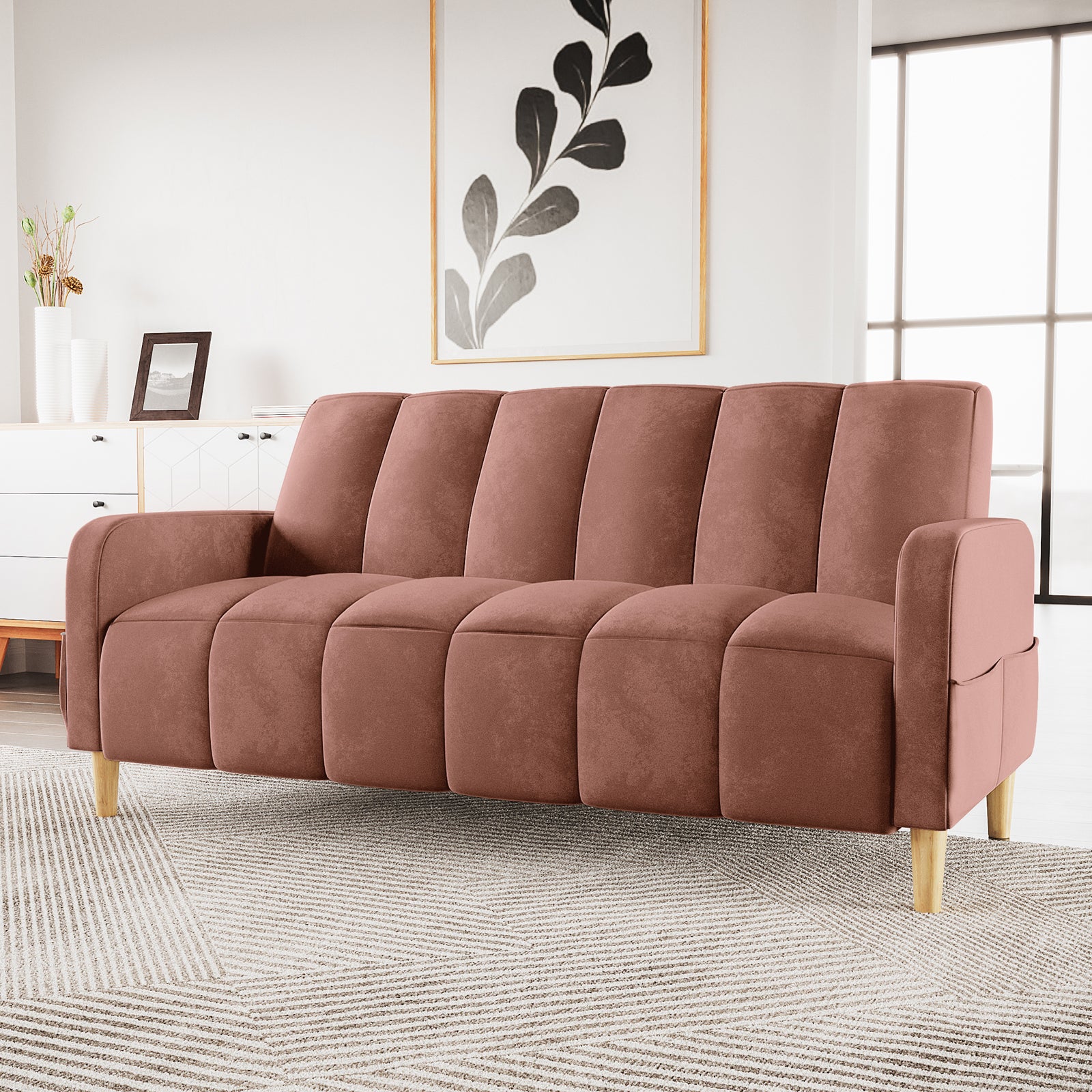 Anica 3 Seater Velvet Sofa Bed in Blush with vertical channel tufting, rose-toned upholstery, and light wood legs, set on a geometric rug in a bright, minimalist room.