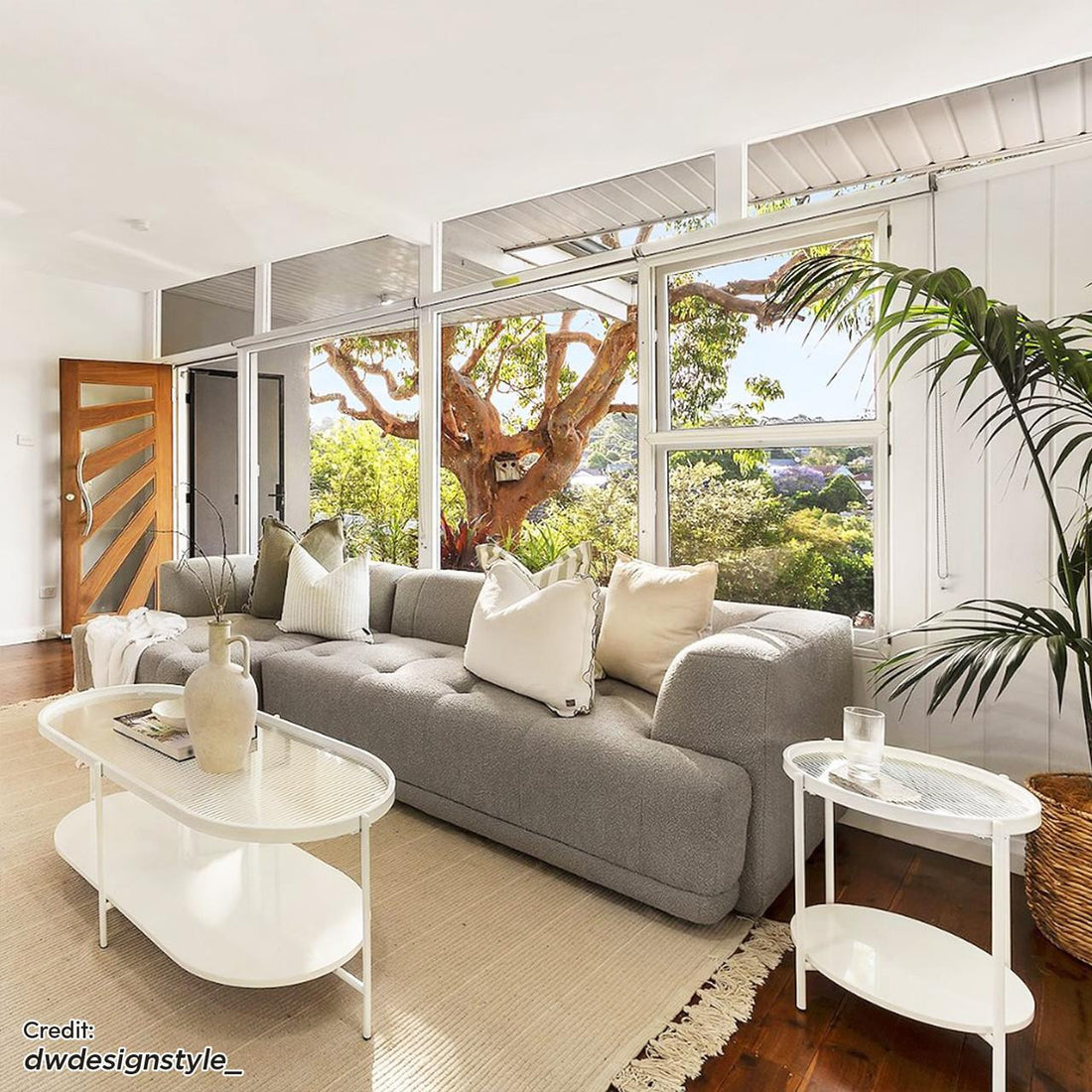 Averill 4 Seater Bouclé Fabric Chaise Sofa in Grey, elegantly tufted and piled with neutral-tone pillows, stands prominently in a bright living room, complemented by a modern coffee table and lush greenery.