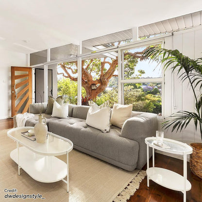 Averill 4 Seater Bouclé Fabric Chaise Sofa in Grey, elegantly tufted and piled with neutral-tone pillows, stands prominently in a bright living room, complemented by a modern coffee table and lush greenery.