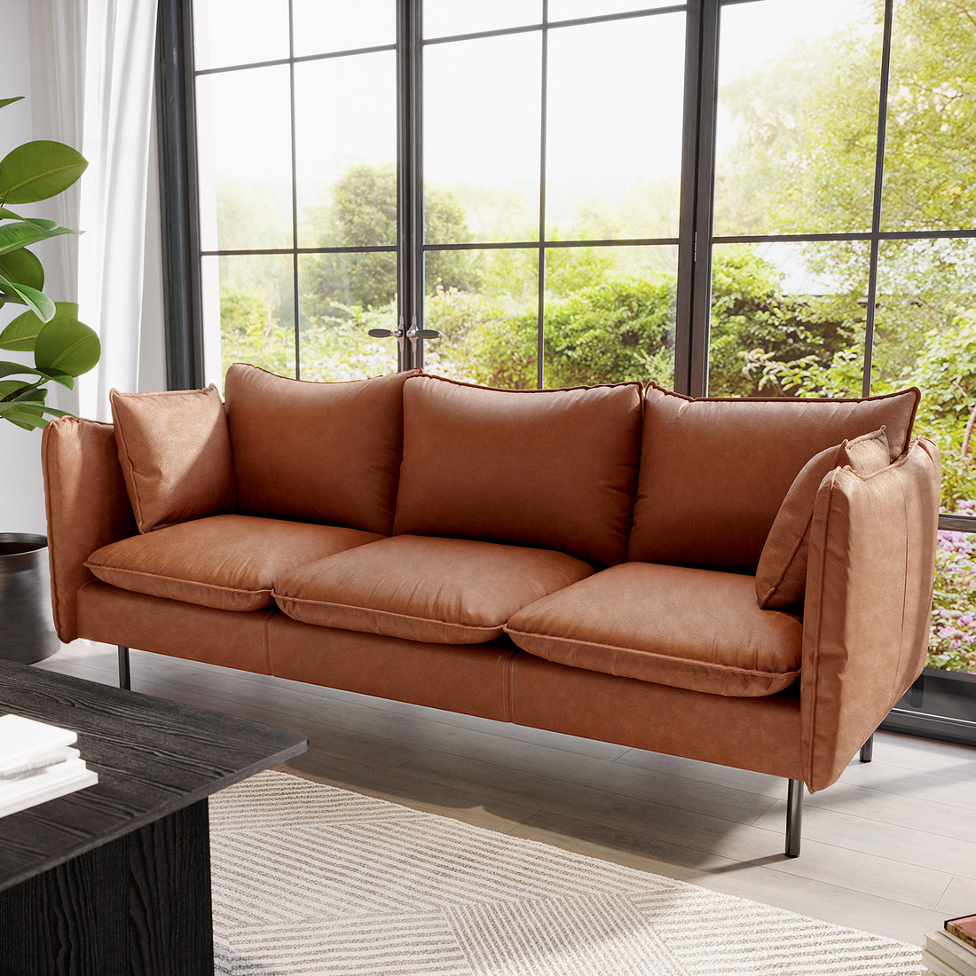 Cassian 3 Seater PU Leather Sofa in Mocha features plush cushions, slender black legs, in a bright living room with large windows and a potted plant, complemented by a dark wood coffee table.