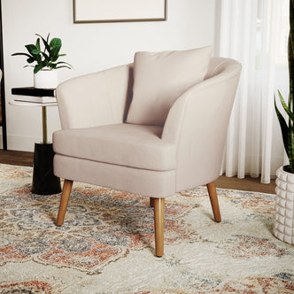 Dalya Linen Accent Chair with Cushion Cream White, featuring blush-pink upholstery, softly curved design, slender wooden legs, and accompanied by a side table with books and a plant, against a minimalist backdrop.