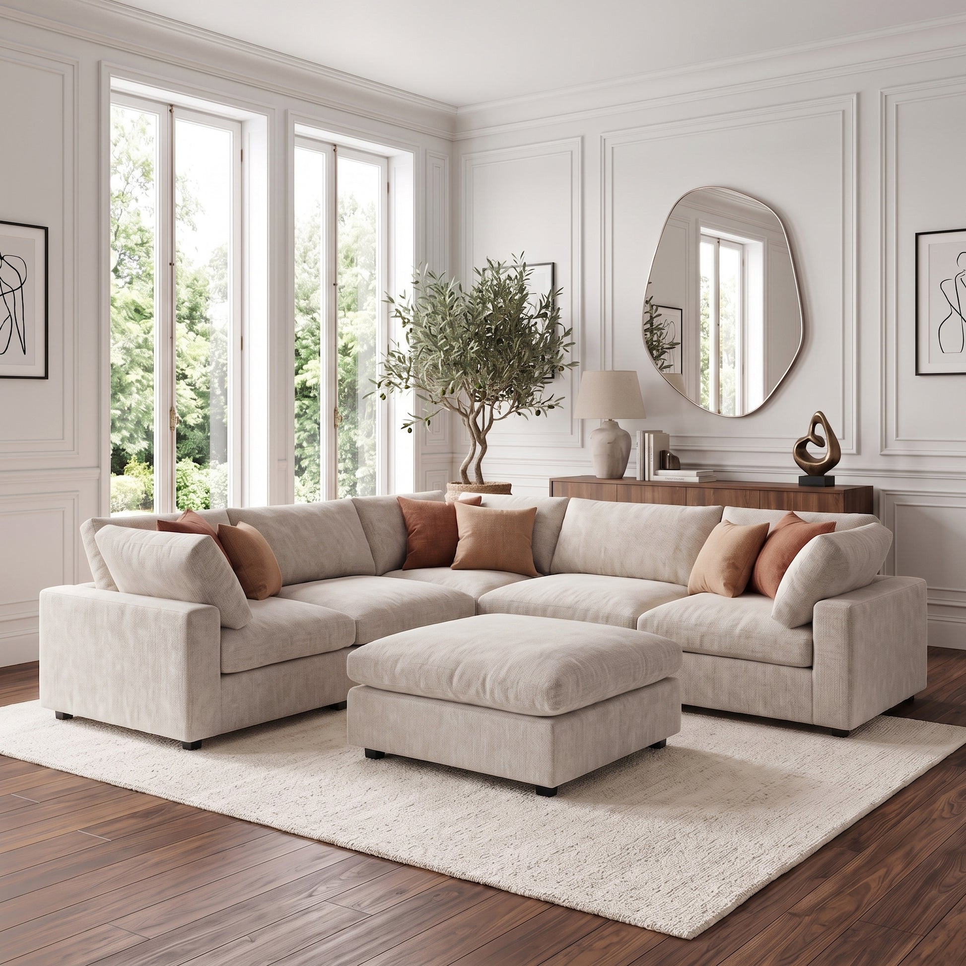 A modern living room featuring a plush, light-colored modular corner sofa with a matching ottoman, styled with rust-colored throw pillows, a large irregular-shaped mirror, and a potted tree on a dark wooden floor.