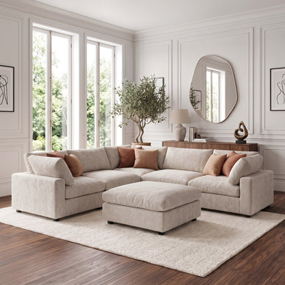 A modern living room featuring a plush, light-colored modular corner sofa with a matching ottoman, styled with rust-colored throw pillows, a large irregular-shaped mirror, and a potted tree on a dark wooden floor.