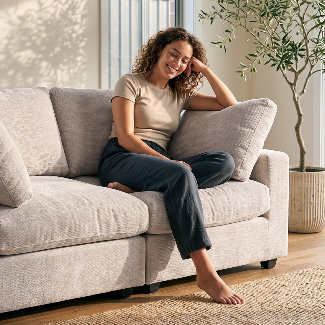 A smiling woman sitting comfortably on a plush, light-colored modular sofa in a bright, sunlit living room with a potted olive tree nearby.